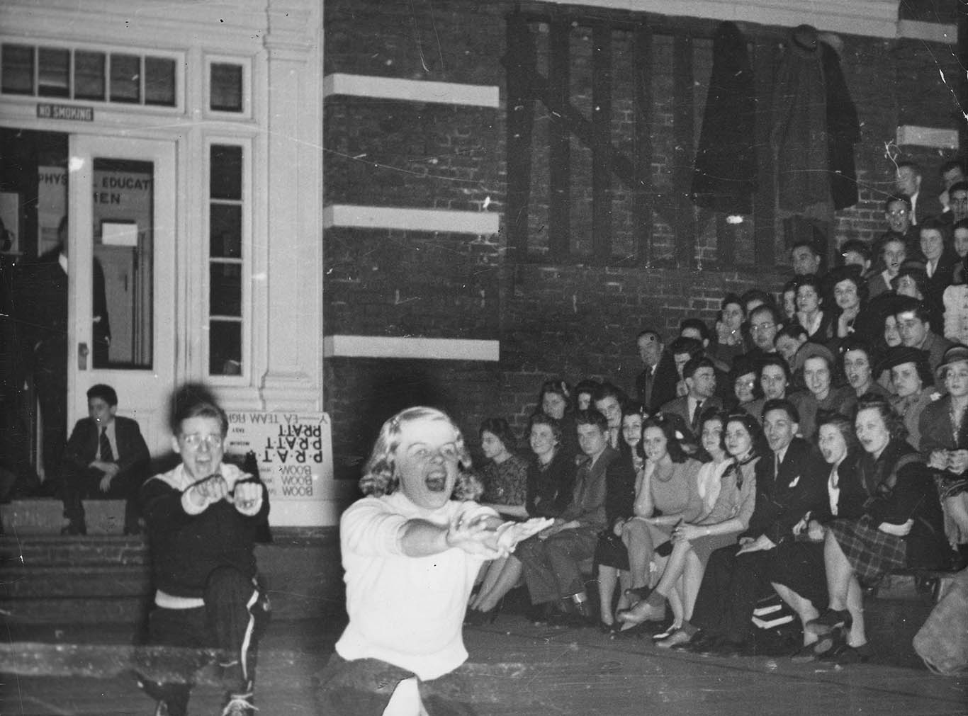 a black and white image of two people walking arms outstretched in front of a seated audience